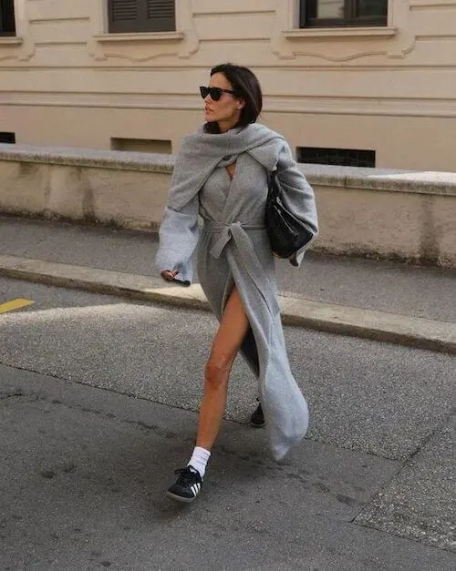 a woman walking wearing a long gray coat and a pair of black Adidas Samba sneakers