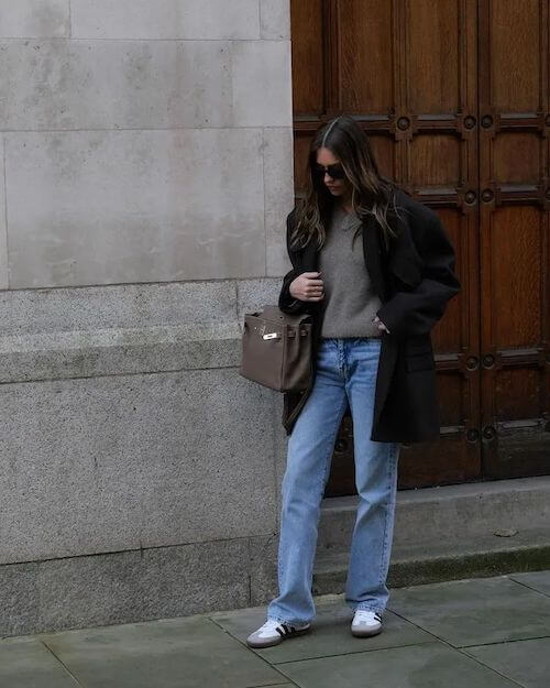 a woman wearing an oversized black blazer, gray knit top, blue jeans, and white Adidas Samba sneakers