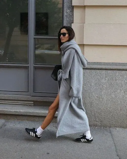 a woman wearing gray maxi coat, gray scarf, and black Adidas Samba sneakers