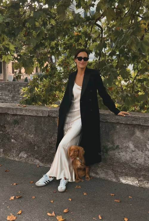 a woman wearing black long coat, and champagne maxi silky dress, and white Adidas Samba sneakers