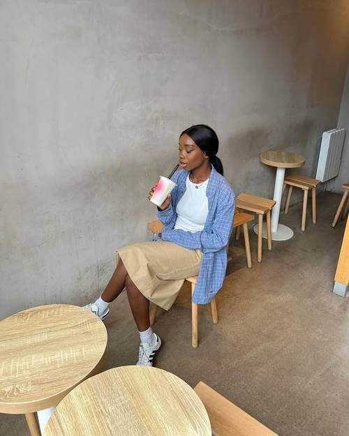 a black woman wearing blue button up shirt, white shirt, beige midi skirt, and white Adidas Samba sneakers