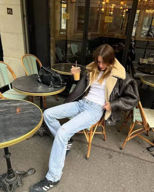 a woman sitting on a chair, wearing a dark brown shearling coat, blue jeans, and black Adidas Samba sneakers