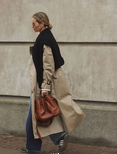 a woman wearing beige trench coat, black sweater, and black Adidas Samba sneakers