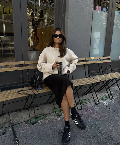 a woman wearing beige oversized sweater, black skirt, and black Adidas Samba sneakers