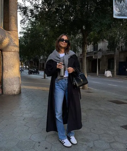 a woman wearing white Adidas Samba sneakers, black coat, white shirt, gray sweater, and blue jeans