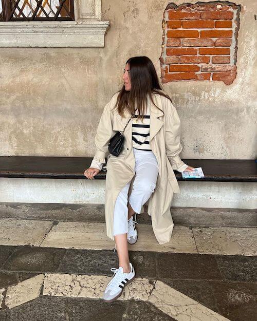 a woman wearing beige trench coat, black and white stripped shirt, white cropped pants, and white Adidas Samba sneakers