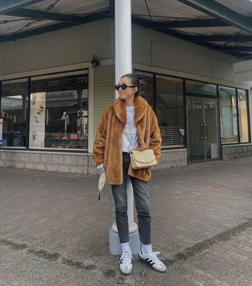 a woman wearing brown faux fur coat, gray skinny jeans, and white Adidas Samba sneakers