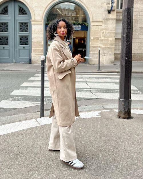 a black woman wearing white Adidas Samba sneakers, beige pants, and beige long coat