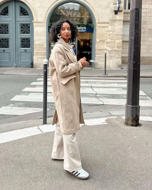 a black woman wearing white Adidas Samba sneakers, beige pants, and beige long coat