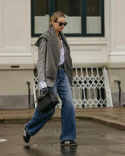 a woman wearing gray blazer, white button down shirt, gray sweater, blue wide leg jeans, and black Adidas Samba sneakers