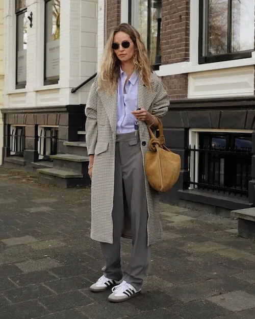 a woman wearing light gray long coat, white button down shirt, gray trousers, and white Adidas Samba sneakers