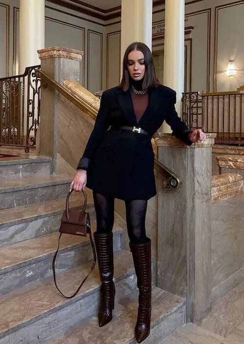 a woman in a luxury hotel wearing a black blazer coat, a luxury brand belt, black tights, sleek knee high boots, and a brown handbang