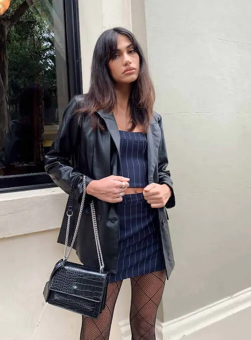 a woman standing in Vegas street, wearing a stylish black leather blazer, navy blue mini skirt set, black tights, and a black handbag