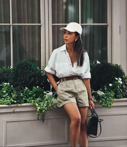 a woman wearing a white button down, a white baseball hat, beige shorts, and a belt