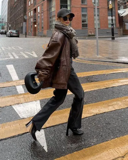 a woman wearing a baseball hat, sunglasses, and brown oversized leather jacket