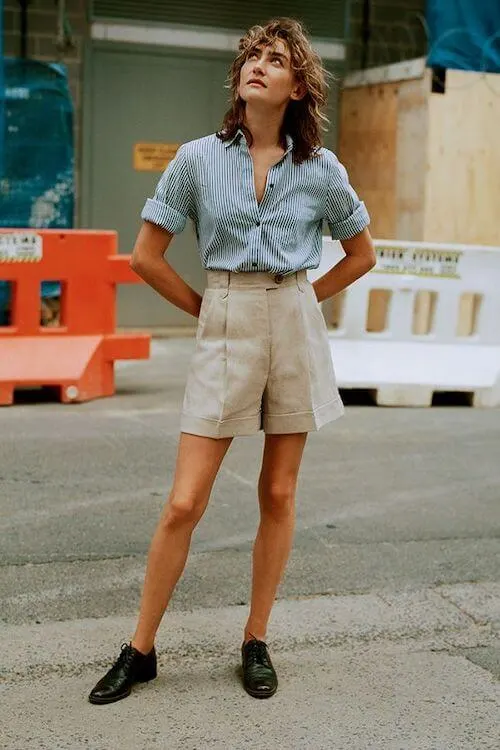 a woman wearing a blue button down shirt, beige shorts, and black shoes