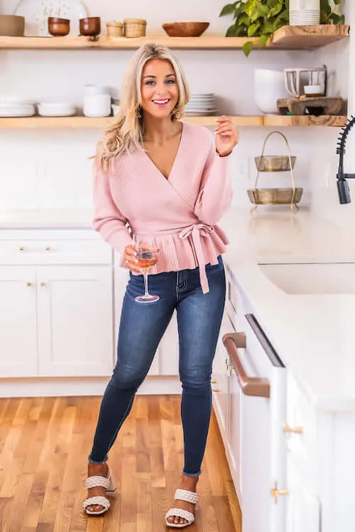 a woman at home wearing a pink wrap sweater top, blue skinny jeans, and beige sandals