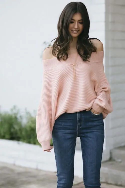 a woman wearing an off the shoulder pink sweater, blue jeans, a long necklace