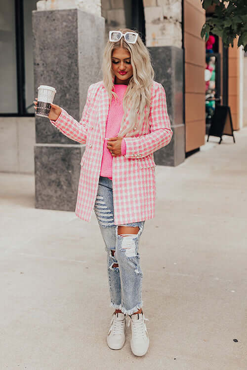 a woman walking in the street wearing a pink knit top, pink blazer, light wash jeans, and white sneakers