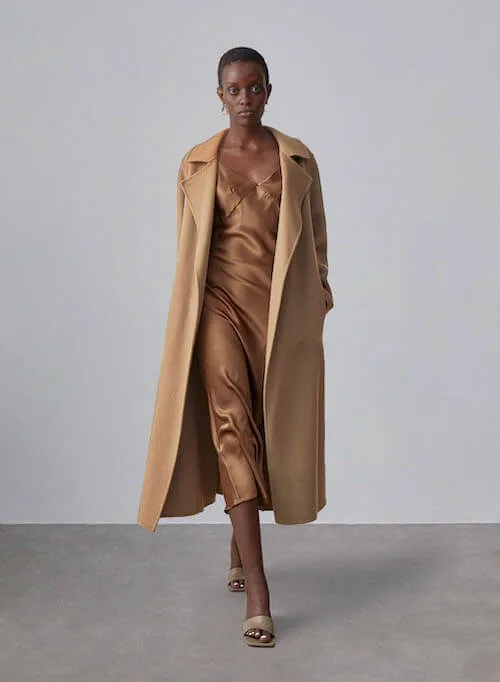 a black woman wearing a camel coat and a long slip dress