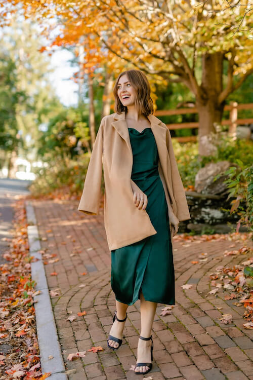 a woman wearing a camel mid-length coat and a green silky dress in the fall