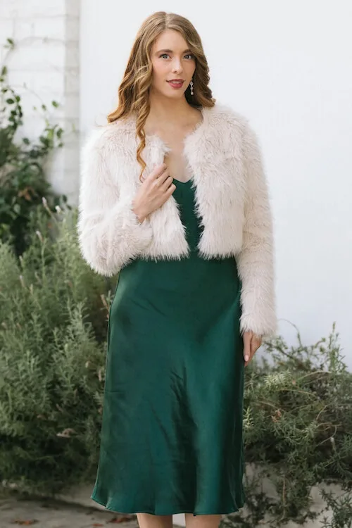 a woman wearing a short cream faux fur jacket and a dark green silky slip dress