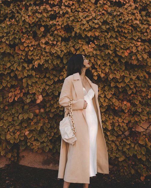 a woman wearing a long camel coat and a white satin slip dress in the fall