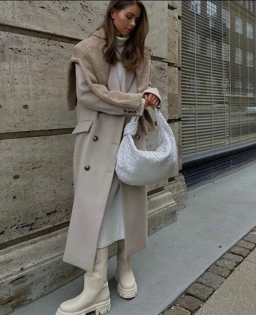 a woman wearing a beige long coat, a white maxi knit dress, a pair of white boots, and with a beige knit top wrapped over her shoulder