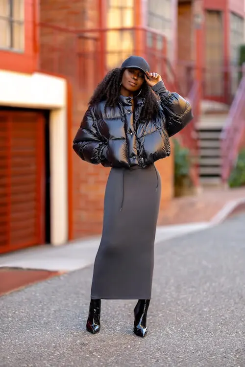 a beautiful black woman wearing a short black puffer jacket, a dark gray maxi dress, a pair of black leather boots, and a dark gray baseball cap