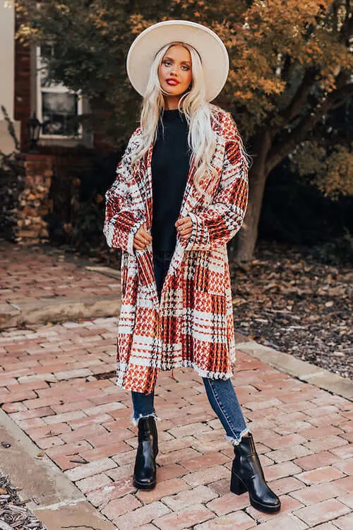 a woman wearing plaid long coat, black sweater, blue skinny jeans, black booties, and a white hat