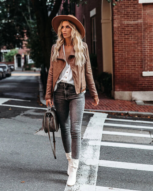 a woman walking in downtown Nashville wearing a brown leather jacket, a white blouse, a pair of dark wash jeans, white ankle boots, and a brown hat