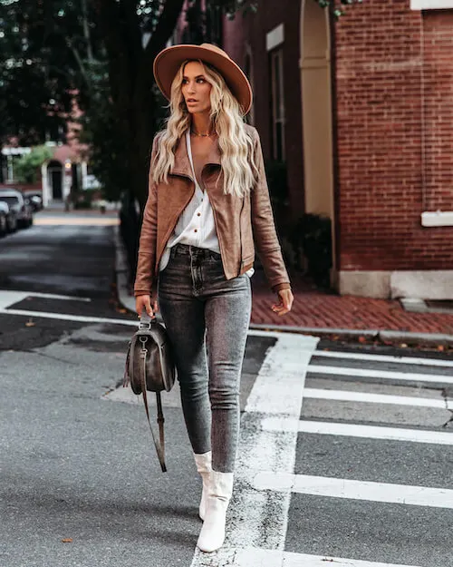 a woman walking in downtown Nashville wearing a brown leather jacket, a white blouse, a pair of dark wash jeans, white ankle boots, and a brown hat