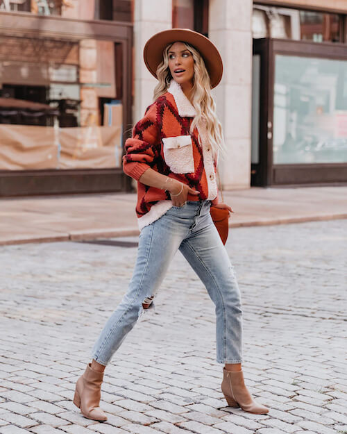 a woman walking ouf of Nashville bar wearing a cozy print cardigan, a pair of jeans, a hat, and a pair of tan ankle boots