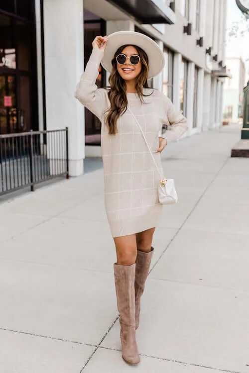 a woman standing on one of Nashville streets, wearing a beige sweater dress, a pair of tan knee high boots, a hat, and a cute white crossbody bag