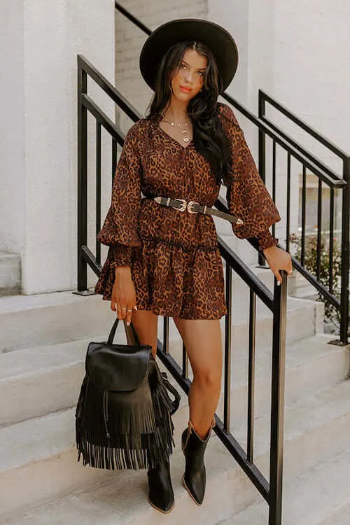 a stylish woman wearing a leopard printed dress, black cowgirl boots, black fringed bag, western belt, and a black hat