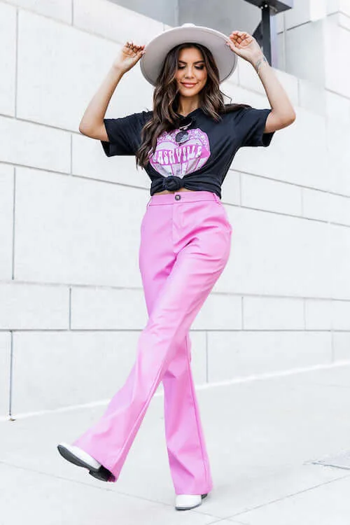 a woman wearing a black graphic Nashville tshirt, a pair of pink leather pants, and a white hat