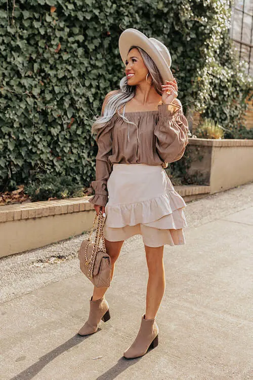 a smiling woman wearing off the shoulder top, white mini skirt, tan ankle boots, and beige hat