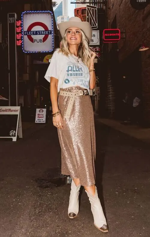 a woman standing in a Nashville street full of bars at night, wearing a letter Tshirt, a glittering midi skirt, a pair of cowgirl boots, and a cowgirl hat